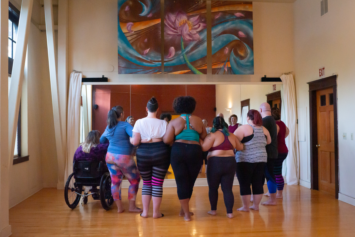 A diverse group of people participates in a dance class in a yoga studio with wooden floors, red walls, and mirrors.