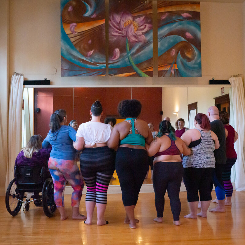 A diverse group of people participates in a dance class in a yoga studio with wooden floors, red walls, and mirrors.